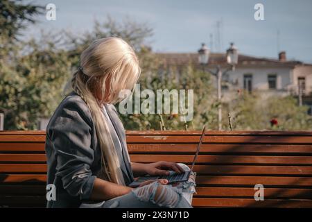 Una donna bionda siede su una panchina con un portatile aperto davanti a lei. Indossa una giacca grigia e ha i capelli in una coda di cavallo. La scena suggerisce un Foto Stock
