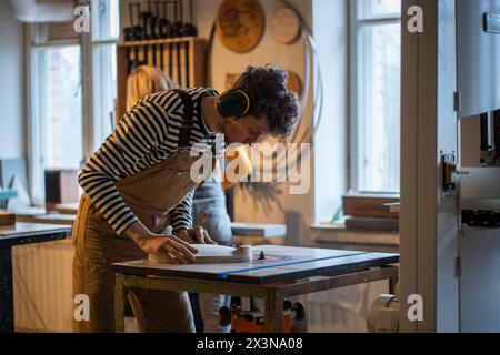 Il falegname uomo concentrato che lavora sul tornio indossando protezioni per le orecchie smeriglia con cura la sedia in legno. Foto Stock