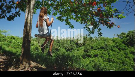 Una donna è seduta su un'altalena appesa a un albero a Bali, in particolare a Ubuds Campuhan Ridge Walk. L'altalena oscilla dolcemente mentre le piace la tranquillità dell'ambiente circostante. Foto Stock