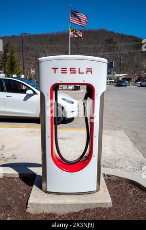 La vale Maryland USA-03 29 2024: A la vale, Maryland, Stati Uniti, un'auto bianca si ricarica presso un Supercharger Tesla sotto la bandiera americana sventolante contro A. Foto Stock