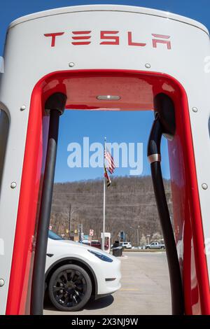 La vale Maryland USA-03 29 2024: A la vale, Maryland, Stati Uniti, un'auto bianca si ricarica presso un Supercharger Tesla sotto la bandiera americana sventolante contro A. Foto Stock