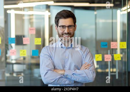 Un uomo con gli occhiali e la barba sorride davanti a un muro ricoperto di note appiccicose. Le note sono gialle e rosa, e sembrano essere correlate a un progetto o a un compito Foto Stock