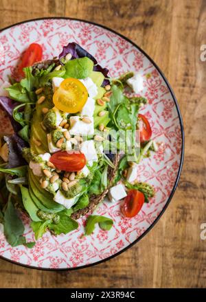 Vista ravvicinata dall'alto dell'avocado con feta, pomodori, pinoli, pesto e insalata su un piatto su un tavolo di legno Foto Stock