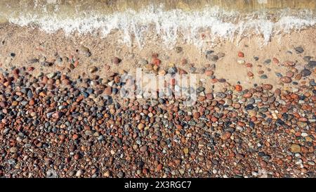 Vista piatta della riva con onde di mare spumeggianti e ciottoli rossi marroni lucidi. Foto Stock