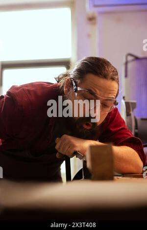Scultore che soffia segatura dal banco di lavoro dopo aver modellato il legno grezzo con scalpello e martello, creando arte del legno. Artista che realizza sculture in legno fatte a mano, realizzando scanalature precise nel blocco di legname con utensili Foto Stock