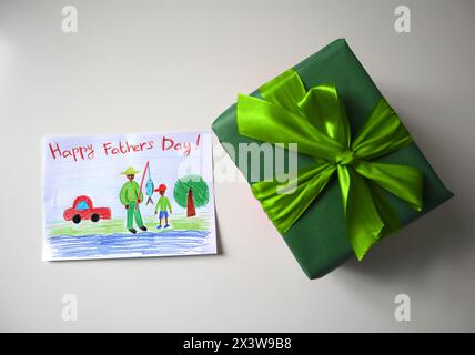 Festa del papà foto di una cartolina disegnata da un bambino. Un regalo di colore verde con un fiocco. Sfondo bianco. La cartolina mostra un padre e un figlio a pescare. Aspetto africano. Disegno con matite colorate. Foto Stock