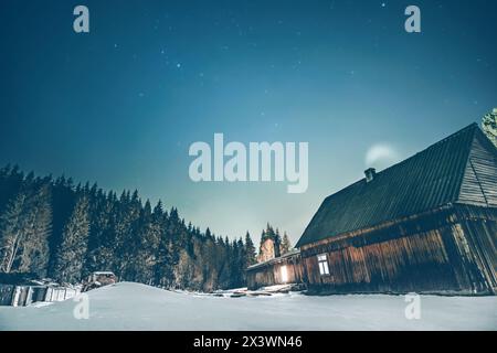 Cottage rurale in legno sulle montagne con finestre che si illuminano al buio durante la notte d'inverno. Capanna in legno sulle colline. Foto Stock