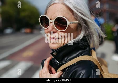 Ritratto di una donna matura elegante con i capelli grigi sulla strada della città. Donna anziana con occhiali da sole in attesa dei trasporti pubblici. Foto Stock