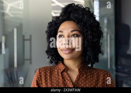 Ritratto di una giovane donna afroamericana sicura di sé con una camicia a pois, che guarda con un look premuroso in un moderno ambiente da ufficio. Foto Stock