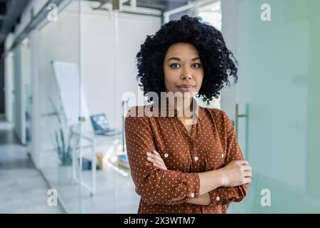 Una giovane donna afroamericana in una camicia a pois si erge con sicurezza con le braccia incrociate in un ambiente elegante, trasmettendo professionalità e sicurezza. Foto Stock