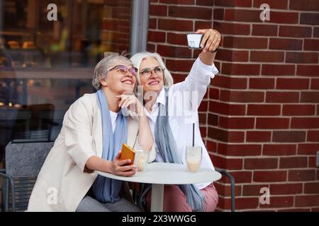 Siede all'aperto Cafe eleganti donne mature con sorrisi che catturano insieme momenti di selfie gioiosi, indossando occhiali e abbigliamento casual elegante, con bevande Foto Stock