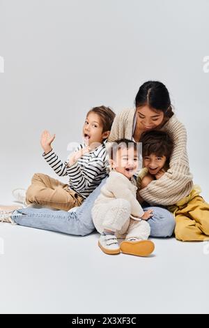 Una giovane madre asiatica seduta a terra circondata dai suoi figli in un ambiente studio su uno sfondo grigio. Foto Stock