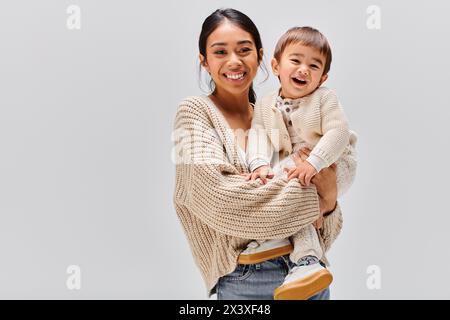 Una giovane madre asiatica tiene tenero il suo bambino tra le braccia in uno studio su uno sfondo grigio. Foto Stock