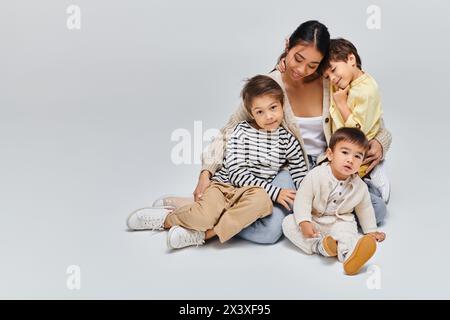 Una giovane madre asiatica siede per terra con i suoi figli in un tranquillo studio su uno sfondo grigio. Foto Stock
