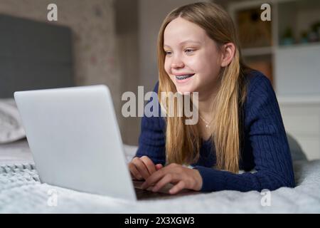 Adolescente che indossa apparecchi ortodontici con laptop a letto a casa, a film o spettacoli in streaming, a navigare online guardando i social media Foto Stock