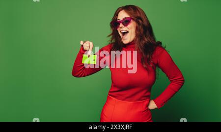 Happy Woman in occhiali da sole alla moda si trova in un vivace studio verde, con una carta di credito. Il suo abbigliamento casual e il suo vivace sorriso incarnano ecologia Foto Stock