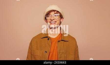 Un giovane allegro con un elegante stile urbano si staglia su un vivace sfondo di pesca. Indossa denim casual, un berretto e occhiali alla moda, sorridendo Foto Stock