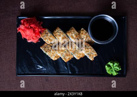 Sushi panino fritto con bacchette e salsa di soia su un piatto nero, realizzato con riso, alghe, ripieni e marrone dorato. Foto Stock