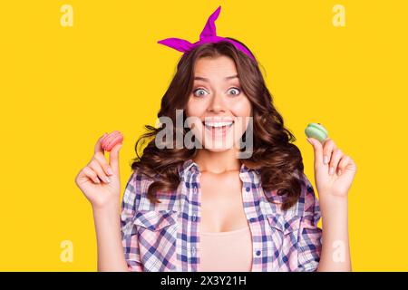 Ritratto di una bella ragazza stupita, pazza, attraente e divertente, con i capelli ondulati, che si diverte a tenersi per mano mostrando due macaron isolati sopra Foto Stock