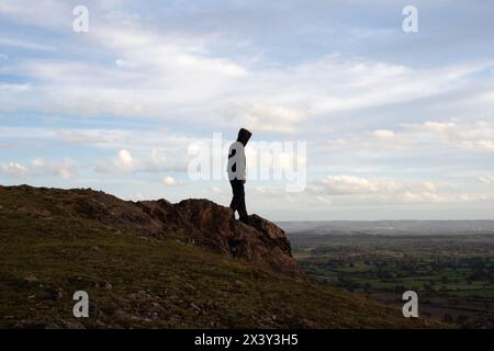 Una figura con cappuccio solitario, in cima a una collina in un'estate che si snoda nella natura. Con terreni agricoli e campagna sullo sfondo. Foto Stock