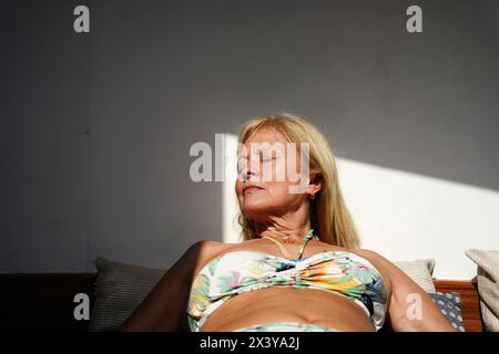 Una donna matura sdraiata su un divano in bikini che prende il sole durante le vacanze estive Foto Stock
