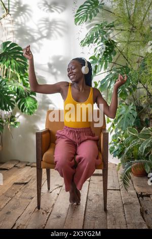 Gioiosa donna nera che balla seduto su una sedia ascoltando la musica preferita circondano piante d'appartamento esotiche. Foto Stock