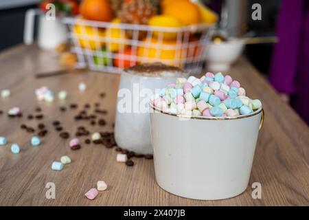 Due tazze su un tavolo di legno, una con cioccolata calda condita con marshmallow, l'altra con caffè, in un ambiente familiare informale con un cesto di frutta Foto Stock