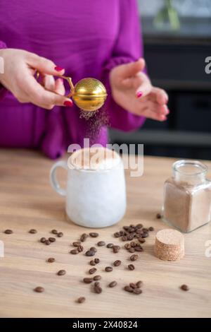 Una mano in un vivace top viola spolvera il cacao su un cappuccino schiumoso, chicchi di caffè sparsi in giro, che incarna il tocco del barista a casa. Pol. Di alta qualità Foto Stock