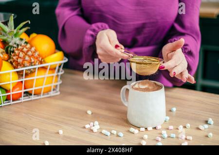 Una persona in una camicetta viola setaccia sapientemente la cioccolata con una bevanda calda, con un vivace cesto di frutta sullo sfondo, creando una mattinata perfetta. Alto Foto Stock