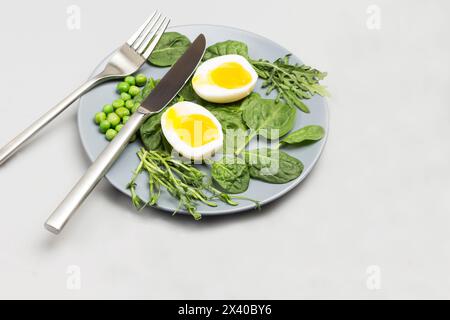 Due metà di uovo di pollo bollito, foglie di spinaci, rucola e piselli verdi su un piatto di ceramica grigia. Forca e coltello sulla piastra. Tovagliolo grigio sul tavolo. Vie superiori Foto Stock