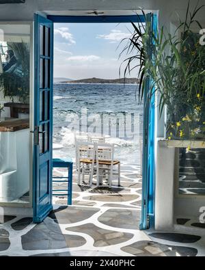 Le porte blu si aprono completamente sull'aria dell'oceano e sulla luce del sole, con il surf che si getta su un patio con sedie; Mykonos, Grecia Foto Stock