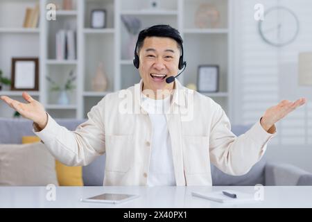 Uomo asiatico di mezza età che indossa la cuffia, sorridente e gestante e offre un servizio clienti da un ambiente accogliente e familiare. Foto Stock