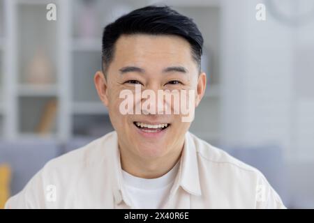 Ritratto di un allegro uomo asiatico di mezza età con un sorriso radioso, che indossa una camicia leggera, seduto in un luminoso e moderno soggiorno. Foto Stock