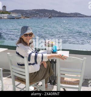 Una donna che indossa un cappello da sole e un paio di occhiali da sole gode di un caffè sul lungomare in una giornata di sole a Mykonos, Grecia; Mykonos, Grecia Foto Stock