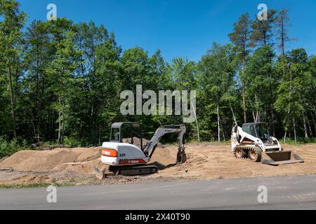 CROW WING CO, MN - 6 giugno 2021: Un miniescavatore Bobcat e caricatore frontale, con cingoli minipala gommata, si trova sullo sporco in un lotto di edifici vuoto fiancheggiato da alberi, in p Foto Stock