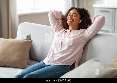 Rilassata donna afroamericana che si diverte con la musica sul divano a casa Foto Stock