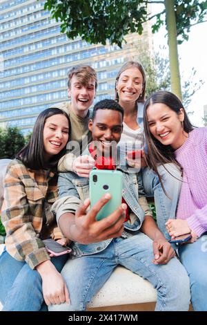 Verticale. Gruppo multirazziale di amici che si divertono e sorridono utilizzando un'app per cellulare. Adolescenti diversi che si divertono a guardare contenuti sui social media con uno smartphone all'aperto. Giovani con il telefono. Foto di alta qualità Foto Stock