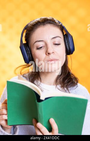 Donna che si concentra sulla lettura di libri, sul relax e sull'ascolto di rumori bianchi, isolata sullo sfondo dello studio. Geek con il romanzo in mano ascoltando canzoni attraverso le cuffie, concetto di intrattenimento Foto Stock