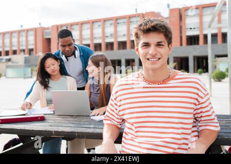 Studentessa che guarda davanti dietro i suoi amici multirazziali mentre usano un laptop. Giovane studentessa caucasica che sorride e guarda la macchina fotografica seduta con i suoi compagni di classe nel campus universitario. Foto di alta qualità Foto Stock