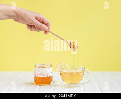 Fai gocciolare il miele su una tazza di tè alle erbe circondato da fiori e bottiglie. Concetto di stile di vita sano e rimedi naturali Foto Stock