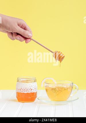 Fai gocciolare il miele su una tazza di tè alle erbe circondato da fiori e bottiglie. Concetto di stile di vita sano e rimedi naturali Foto Stock