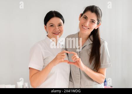 Cosmetologi femminili felici in uniforme che modellano il cuore con le dita e sorridono alla macchina fotografica. Bellissimi e giovani dermatologi che lavorano insieme in una clinica di bellezza. Foto Stock