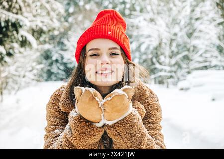 Giovane donna con abiti invernali marroni e cappello rosso che soffia fiocchi di neve, che si tiene nelle sue mani nella foresta invernale. Atmosfera invernale Foto Stock