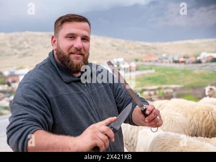 Un macellaio che tiene in mano un coltello, con la lama del coltello che si interseca, come se lo affilasse per tagliare le pecore durante la festa, con una bella, Foto Stock
