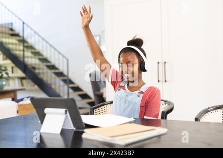 La ragazza afroamericana con le cuffie alza la mano in una lezione online basata su tablet Foto Stock