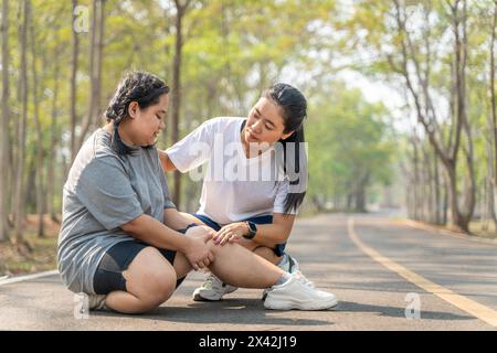 Donna in sovrappeso con lesioni al ginocchio seduta sulla pista da corsa in un parco di corsa tenendo il ginocchio doloroso mentre la sua amica di corsa seduta besi Foto Stock