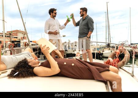 Due uomini si impegnano in una conversazione amichevole con bevande mentre le donne leggono libri, tutti godendosi un pomeriggio rilassato su uno yacht al porticciolo. Foto Stock