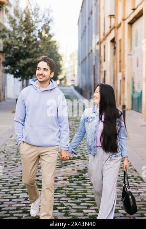 Coppia interrazziale che cammina su un marciapiede tenendosi per mano. Giovane uomo e donna che camminano per la strada della città. Foto Stock