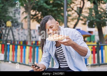 Giovane africano che mangia pizza all'aperto. Uomo che mette tutta una fetta di pizza in bocca mentre è seduto fuori usando il suo smartphone. Foto Stock