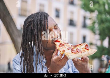 Giovane africano di mangiare pizza slice all'esterno. L'uomo mettendo tutta la fetta di pizza nella sua bocca mentre è seduto all'esterno. Foto Stock